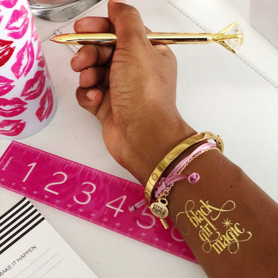 woman's hand with bracelets holding a gold pen on table top with pink ruler and pink lipstick mug.