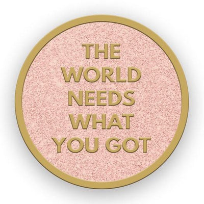 enamel pin with “The World Needs What You Got” in gold letters on a pink background