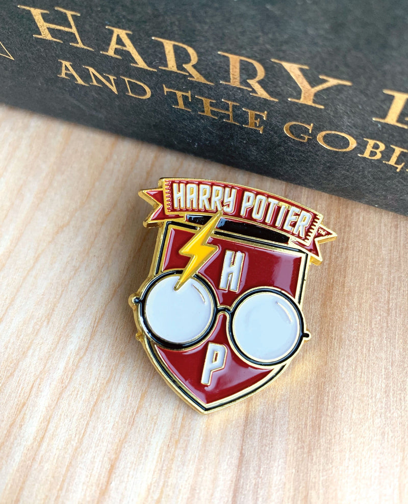 Harry Potter Glasses enamel pin shown on a wood surface next to an upright book with the words HARRY... AND THE GOBL...