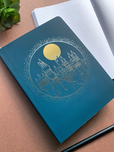 Harry Potter Hogwarts softcover journal notebook shown laying on a desk. Journal cover features a Hogwarts castle illustration printed in silver foil on a blue  background.