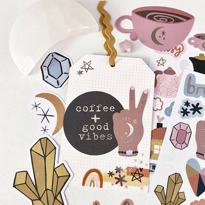 Handmade gift tag on white grid background, brown die cut circle with words "coffee + good vibes", die cut illustration of hand and fingers doing a peace sign. Elements from the Mandy Ford Soul Care craft kit scattered in the background.