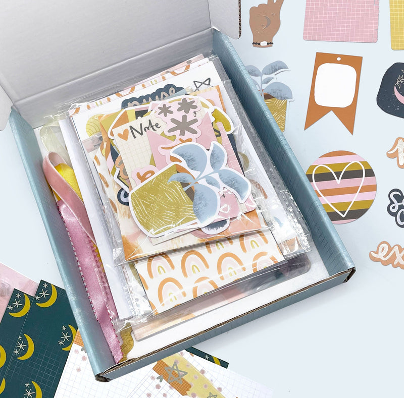craft kit components including stickers and die cuts shown in a box featuring navy, gold and pink patterns and illustrations shown on a blue background.