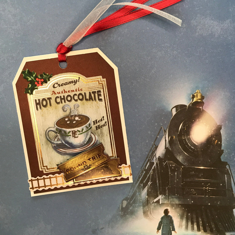 craft kit featuring a hot chocolate gift tag shown with red ribbon on top of a scrapbook paper of the Polar Express from the paper crafting kit.