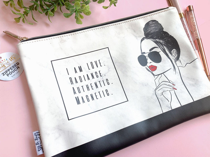 Black and white pencil pouch with gold zipper featuring an illustration of a woman with red lips and words of inspiration shown on a pink background with 2 pens.