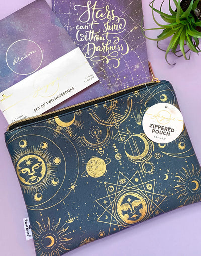 Navy pencil pouch with gold zipper featuring gold celestial illustrations shown on a purple background with a set of two purple notebooks.