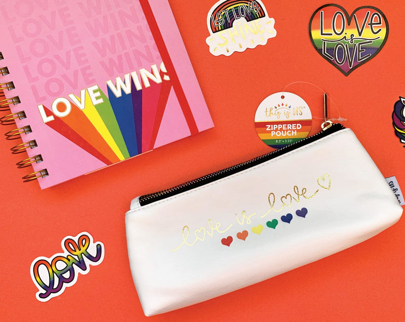 White pencil pouch with gold zipper featuring six hearts in rainbow colors with the words Love is Love in gold script shown on red background with rainbow themed shaped stickers and a pink spiral notebook.