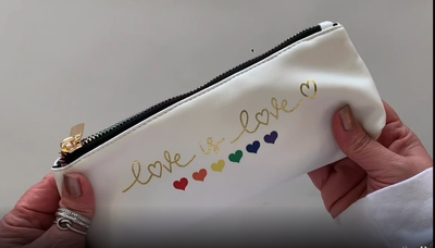 Female hands pick up and open a rainbow, metallic zippered pencil pouch featuring rainbow hearts with a gold script "love is love" on a white background with a black interior filled with various items..