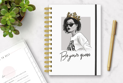 Gold spiral planner organizer features a woman wearing a gold crown and a shirt with the words GIRL POWER partially shown, against a gray and white background. The words Be your queen are printed in black script below. Also features a black elastic page holder.