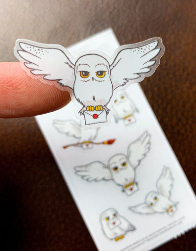 Sheet of Harry Potter stickers featuring illustrations of Hedwig, shown on a white background. One sticker is shown removed from sheet and attached to an index finger to show detail.