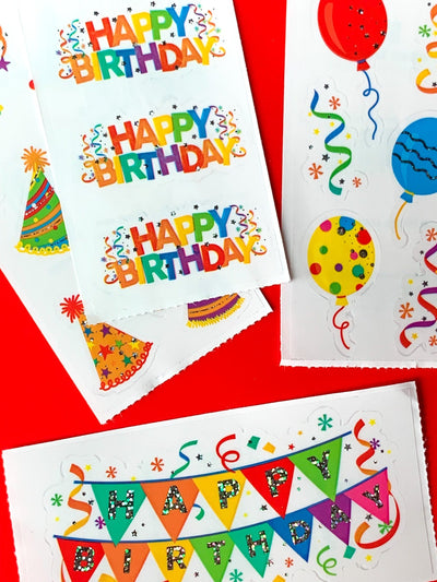 close up of 4 sheets of birthday stickers featuring colorful Happy Birthday banners, balloons and party hats with holographic foil, shown on red background.