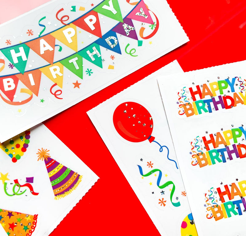 close up of 4 sheets of birthday stickers featuring colorful Happy Birthday banners, balloons and party hats with holographic foil, shown on red background.