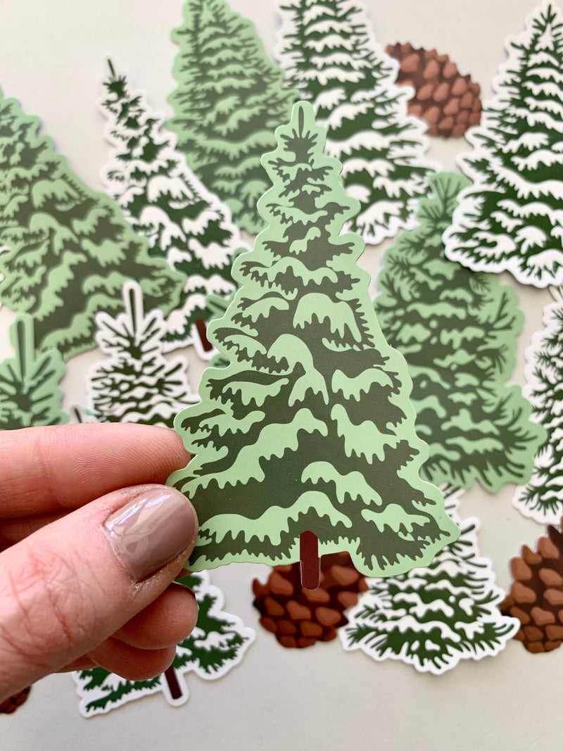 hand displays close up of  illustrated, die cut pine tree sticker, shown in front of an assortment of other pine trees and pine cone scrapbook stickers.