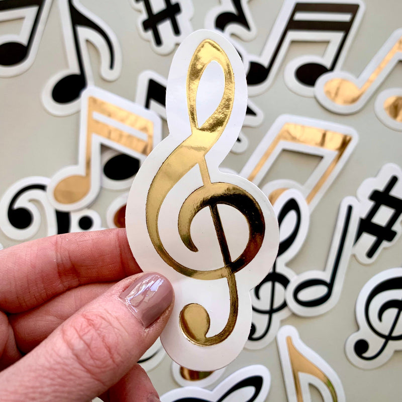 hand displays close up of die cut, gold foil music note scrapbook sticker in front of an assortment of more musical note scrapbook stickers.
