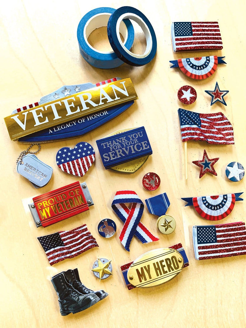 3D scrapbook stickers featuring US flags, and red, white and blue veteran themed sentiments shown next to 2 rolls of washi tape on a wood surface.