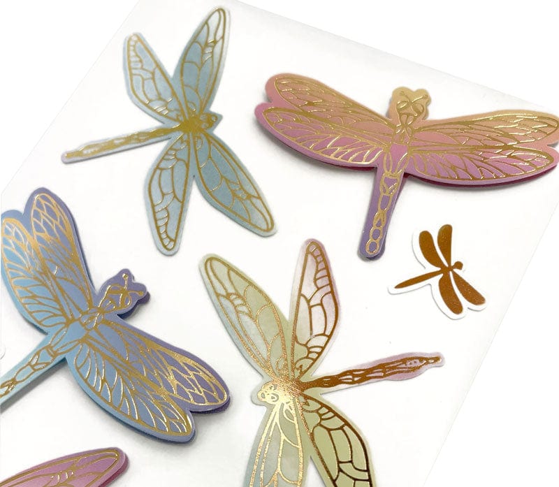 close up of scrapbook stickers featuring pastel colored, illustrated dragonflies with gold details.