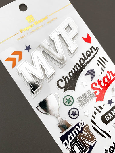 Scrapbook sticker featuring "MVP" and "champion" words. Silver foil details and red, blue and black symbols, shown in white package on gray background.