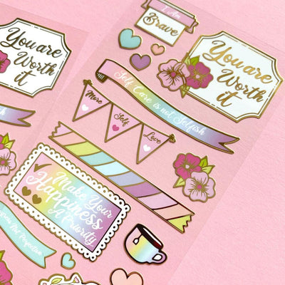 close up of foil stickers featuring illustrated flowers, hearts and self-care illustrations with gold details, shown on pink background.