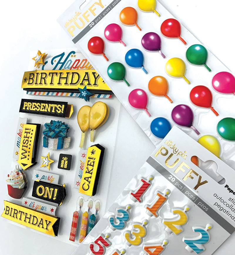 An assortment of happy birthday themed scrapbook stickers featuring words, presents, numbers, candles and balloons with gold foil and clear glitter details shown layered on a white background.