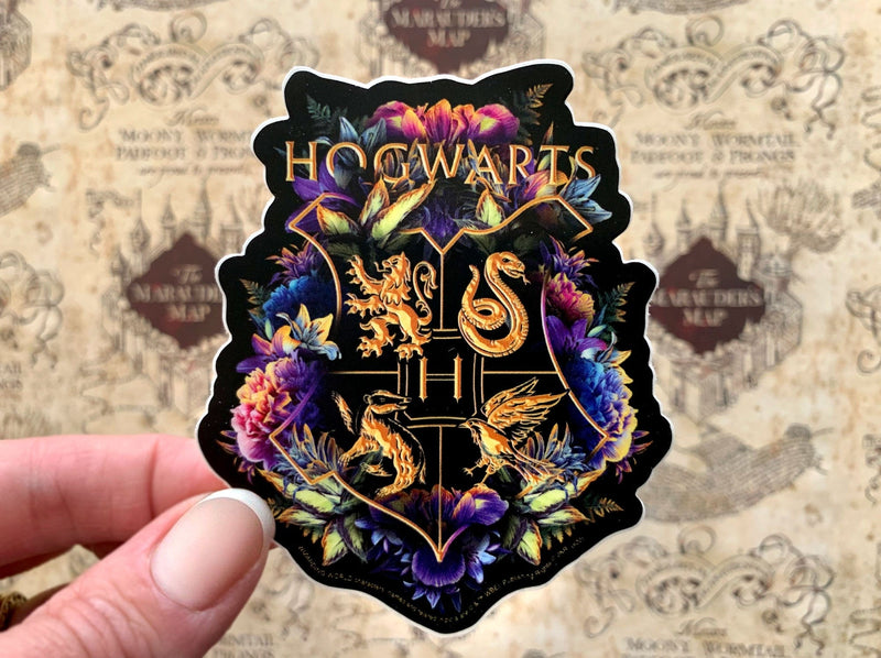 close up of shaped Harry Potter laptop sticker, shown in hand, featuring the Hogwarts crest with floral and black and gold details, shown over a beige pattern of the  marauder&