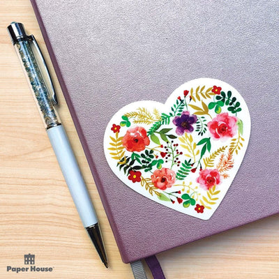 Heart shaped laptop sticker featuring watercolor floral illustrations with gold details, shown on a metallic lavender journal with a white pen beside it.