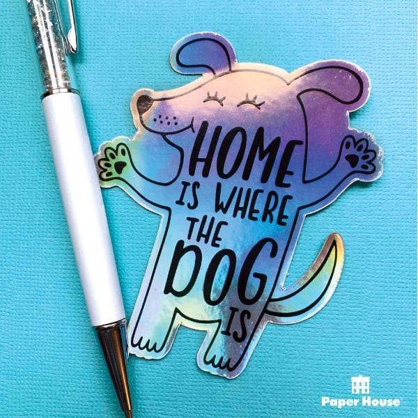 Shaped laptop sticker featuring a holographic illustrated dog with the words "Home is where the Dog is", shown on a blue background with a white pen beside it.