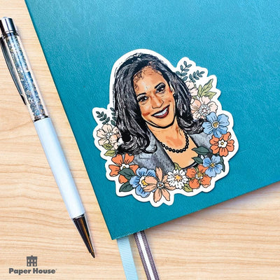 Shaped laptop sticker featuring an illustrated floral portrait of Kamala Harris is shown on a blue notebook with a white pen beside it.