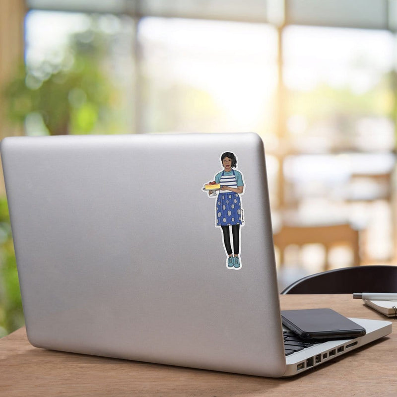 Shaped, vinyl sticker featuring a woman of color dressed in a blue patterned apron, holding a cake on a cake plate shown on a silver laptop.