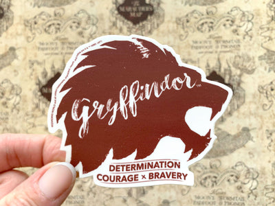 close up of shaped, laptop sticker featuring words Gryffindor, determination, courage and bravery, held in hand over a marauder's map background.