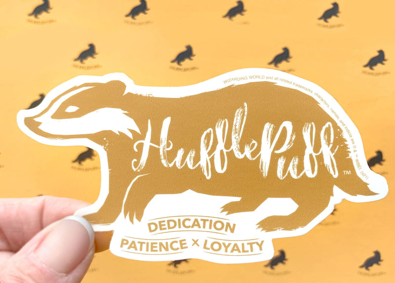 close up of shaped, vinyl sticker featuring a gold skunk with the words Hufflepuff, dedication, patience and loyalty, shown in hand over a pattern of black skunks on yellow background.