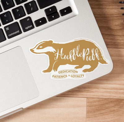 Shaped, vinyl sticker featuring a gold skunk with the words Hufflepuff, dedication, patience and loyalty shown under a laptop keyboard on a wood surface.