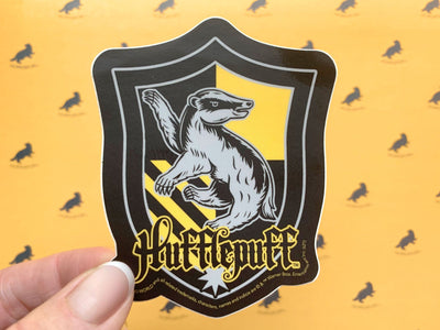close up of vinyl laptop sticker featuring Harry Potter Hufflepuff Shield with yellow and black detail, held in hand shown over pattern of black badgers on yellow background.