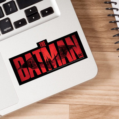 vinyl sticker featuring a black background with red Batman letters and a bat shown on a laptop on a wood surface.