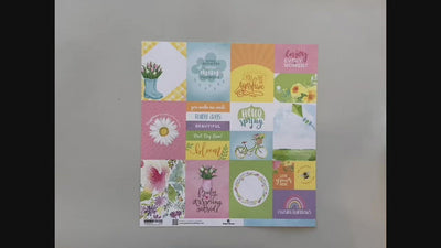 Female hands pick up scrapbook paper featuring pastel spring illustrations and sentiments on one side, and a blue rain pattern on the reverse.