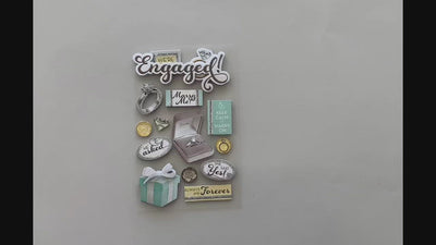 Female hands pick up and show front and back of scrapbook stickers featuring engagement themed items with silver and teal details.
