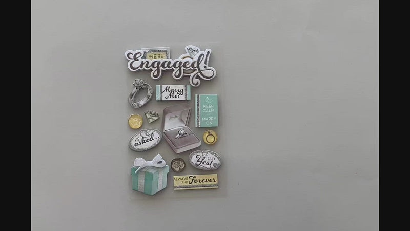 Female hands pick up and show front and back of scrapbook stickers featuring engagement themed items with silver and teal details.