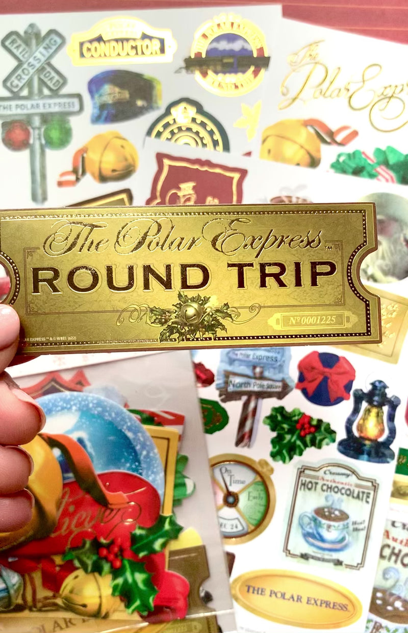 Hand displaying a close up of die cut scrapbook sticker featuring a Polar Express gold ticket  shown over sticker sheets of scenes, bells and tickets with gold details, on red background.