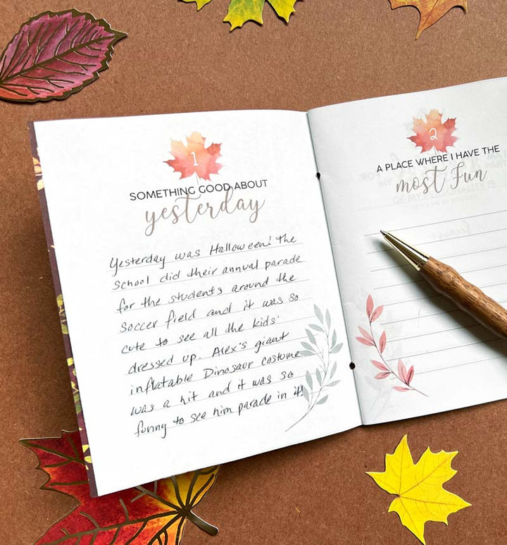 DHT-0002E - Gratitude Journal - Printable - Open gratitude journal with handwritten entries and watercolor fall leaf designs, surrounded by decorative autumn leaves on brown background