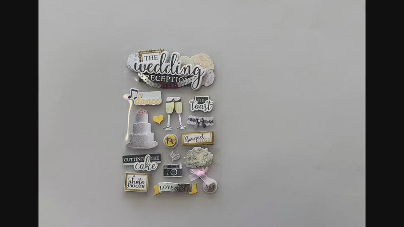Female hands pick up and show front and back in detail of 3D scrapbook stickers featuring wedding cake, champagne and words of love with gold and black details.