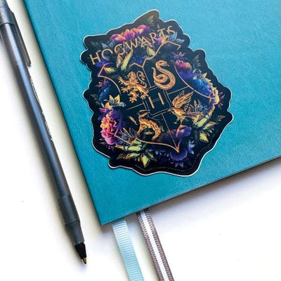Shaped Harry Potter laptop sticker featuring the Hogwarts crest with floral and black and gold details is shown on a blue notebook with a black pen beside it.