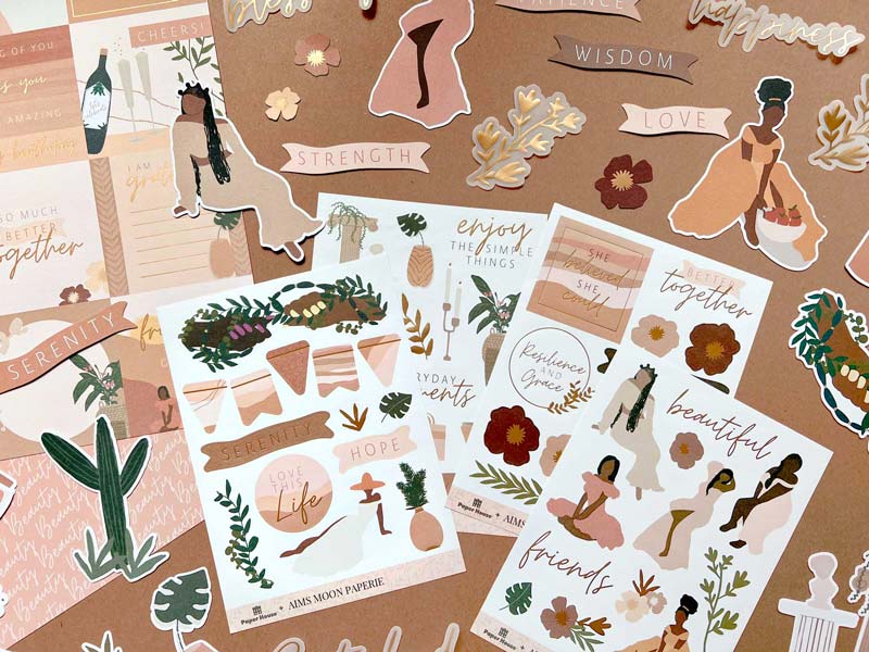 KIT-0129E - Craft Kit - Aim's Moon Paperie - Serenity - Flat lay of sticker sheets and cards in warm earth tones featuring floral designs, illustrated women, botanical elements, and inspirational words like "serenity" and "strength."