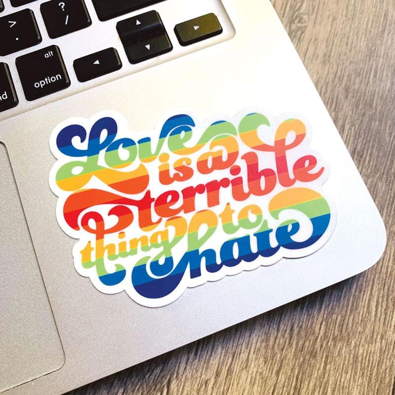 Shaped laptop sticker featuring rainbow colored "Love is a terrible thing to hate" script text, shown on a silver laptop computer.