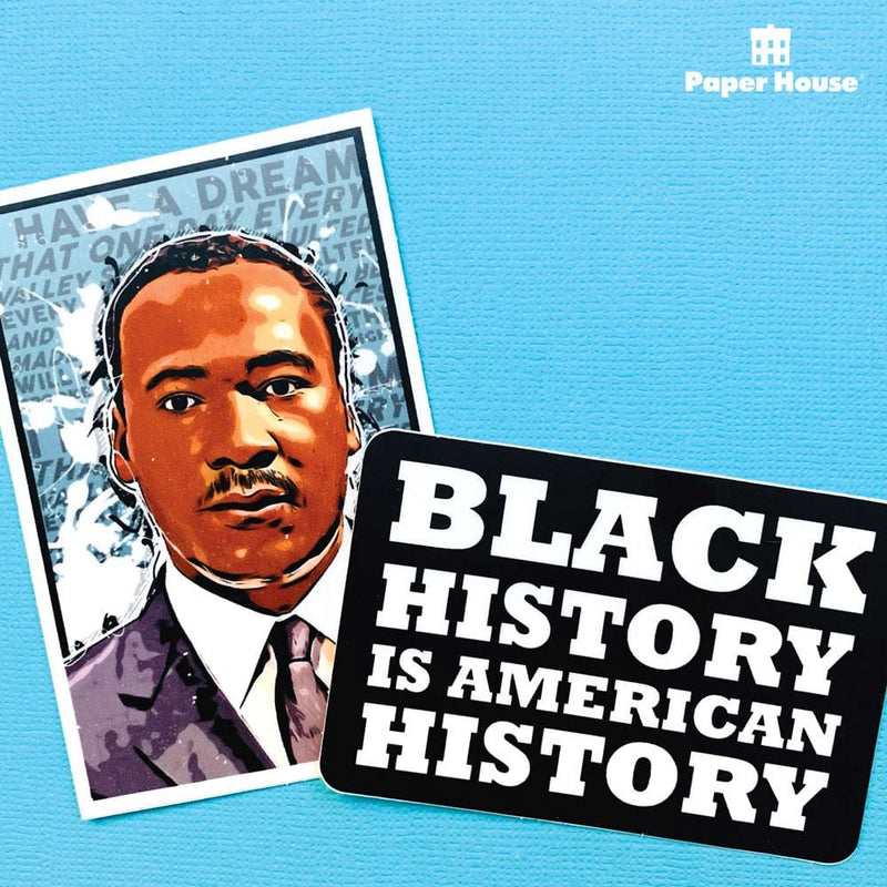 "BLACK HISTORY IS AMERICAN HISTORY" laptop sticker in white letters on a black background, is shown overlapping another laptop sticker featuring Martin Luther King Jr. Both are shown on a blue background.