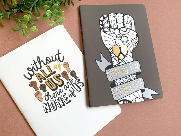 journal notebook set shown overlapping featuring "equality" depicted in black and white graphic illustration with gold details.