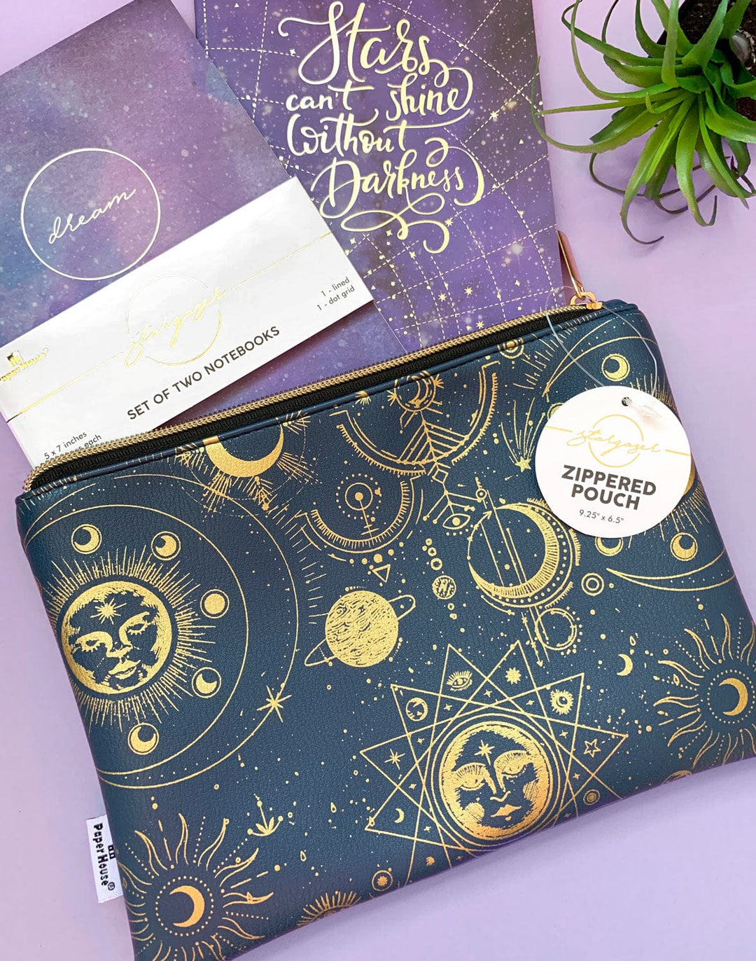 Navy pencil pouch with gold zipper featuring gold celestial illustrations shown on a purple background with a set of two purple notebooks.