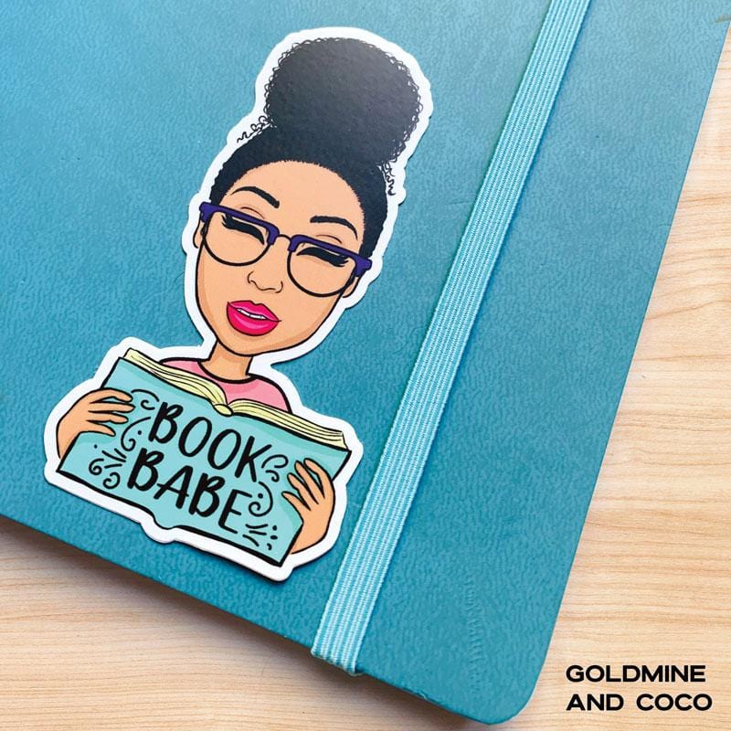 Shaped laptop sticker featuring an illustration of a woman wearing glasses, reading a book that says Book Babe, shown on a blue notebook with elastic band.