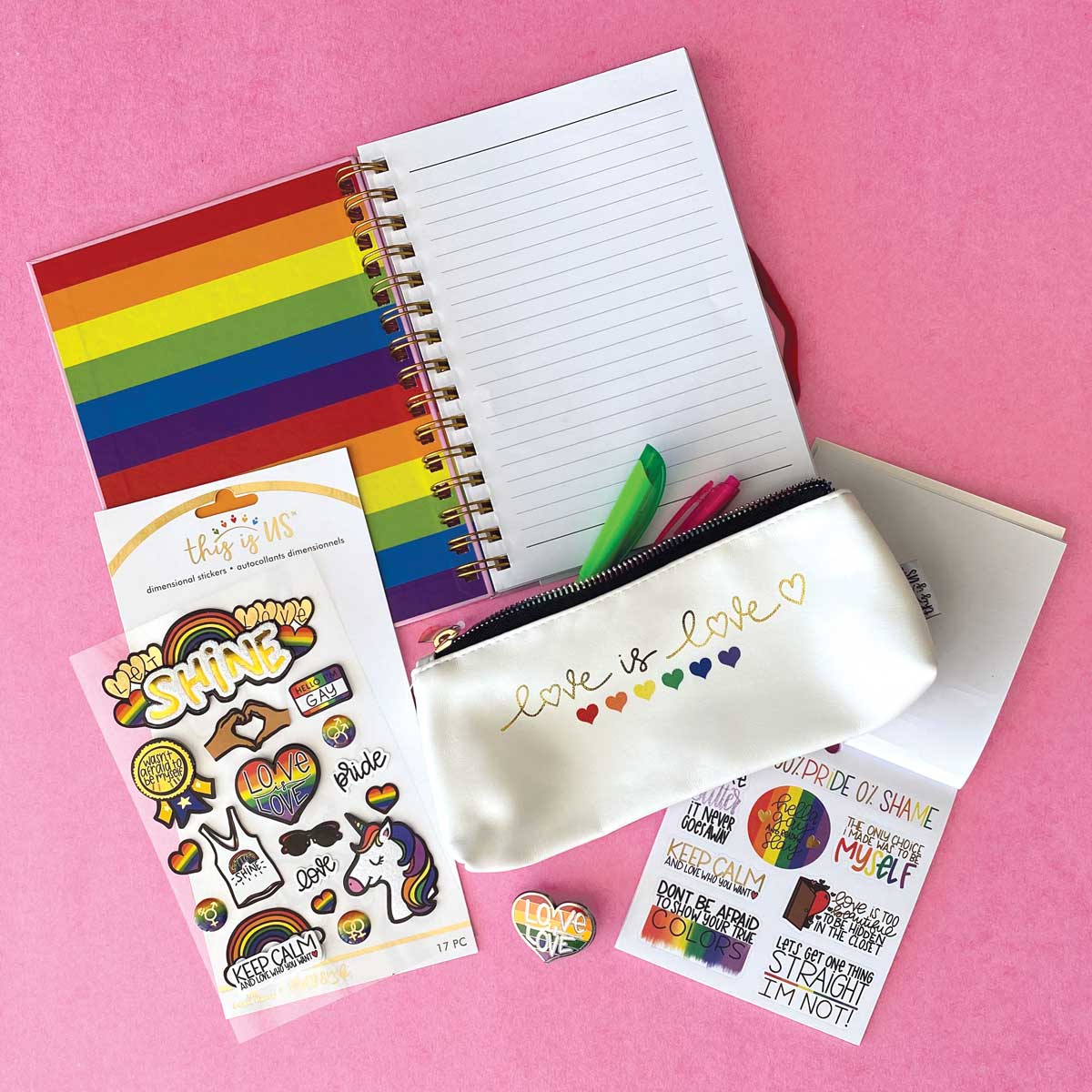 spiral notebook, pencil pouch, stickers and an enamel pin featuring rainbows are shown on a pink background with pens.