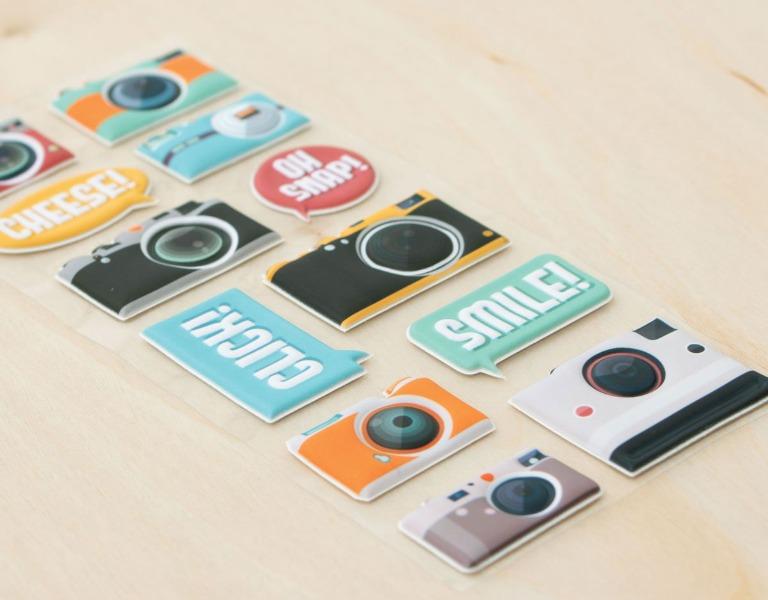 close up of puffy stickers featuring cameras, shown on an angle on a beige surface.