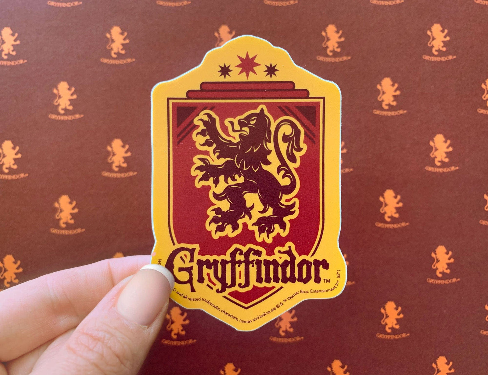 close up of vinyl laptop sticker featuring Harry Potter Gryffindor Shield with red and yellow detail, shown in hand over a gold an red pattern.