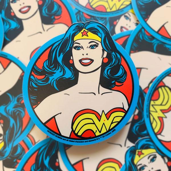 Shaped laptop sticker featuring the Wonder Woman portrait in illustrative style with blue, red and yellow details, shown on top of a large quantity of more Wonder Woman stickers.